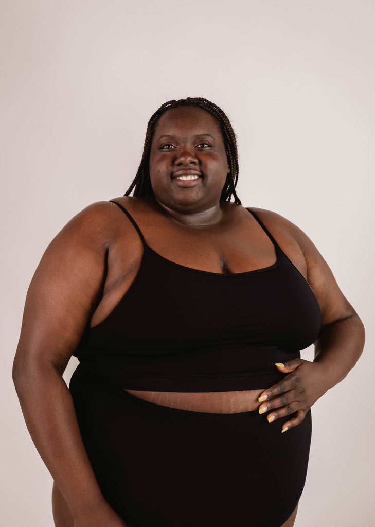 Person stands against a plain background, wearing a Mimi & August Mango Black Bralette Bikini Top with adjustable straps and matching bottoms, smiling with one hand resting on their stomach.