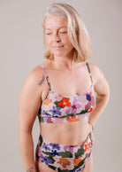 A woman with grey hair, wearing a Mimi & August Mango Botanica Bralette Bikini Top with adjustable straps and leggings, stands with her eyes closed and a slight smile, displaying a tattoo on her left shoulder.