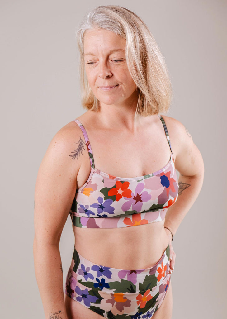 A woman with grey hair, wearing a Mimi & August Mango Botanica Bralette Bikini Top with adjustable straps and leggings, stands with her eyes closed and a slight smile, displaying a tattoo on her left shoulder.