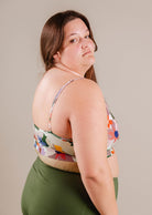 A woman in a Mimi & August Mango Botanica Bralette Bikini Top with adjustable straps and green shorts stands confidently, looking over her shoulder at the camera.