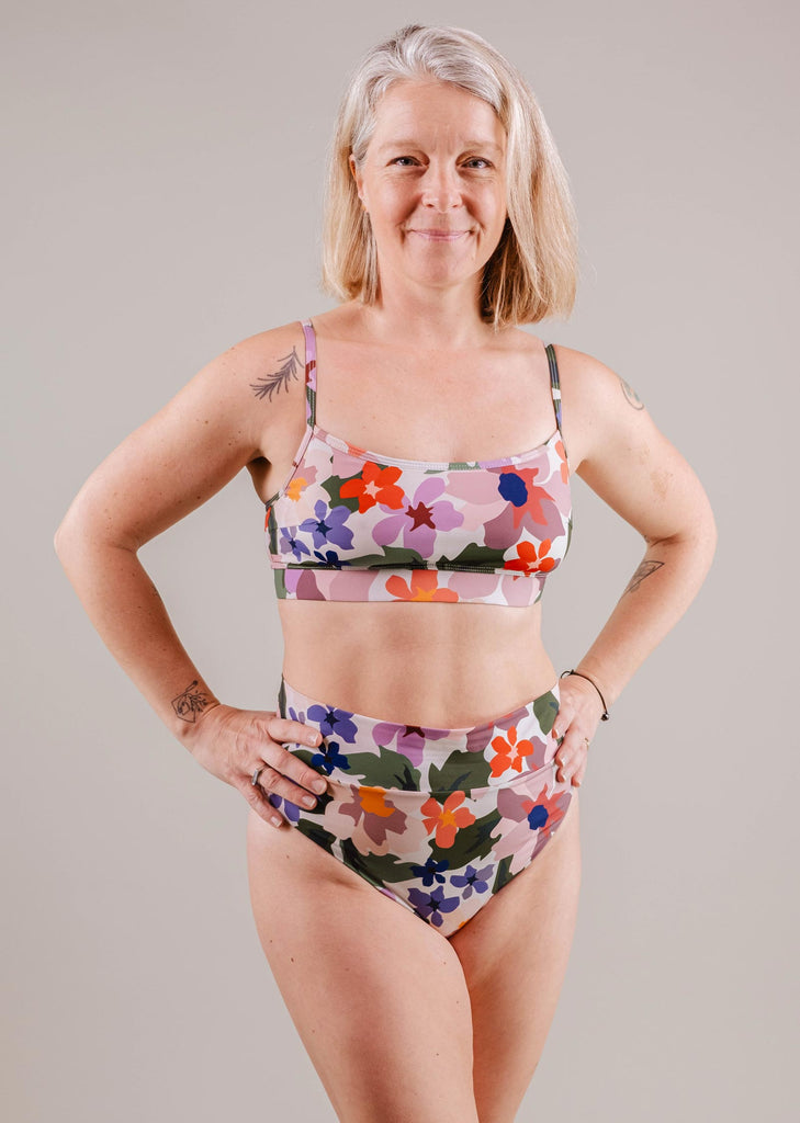 A middle-aged woman with a confident smile wearing a Mimi & August Mango Botanica Bralette bikini top stands with hands on hips against a neutral background.