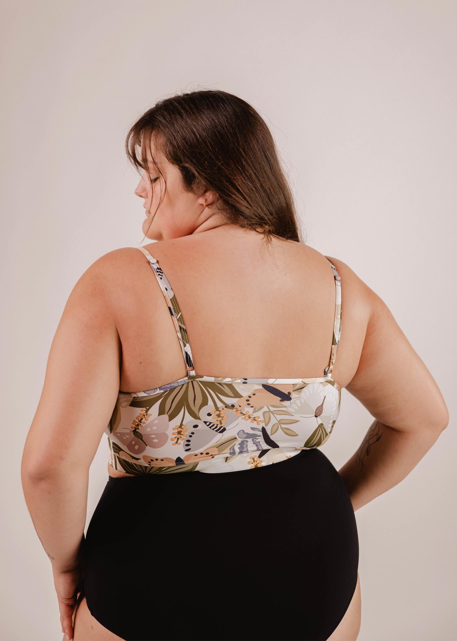 A woman wearing a floral print, sleeveless top and a black skirt or shorts, viewed from the back. The vibrant butterfly patterns on her Mimi & August Mango Mariposa Bralette Bikini Top provide excellent support while adding a touch of elegance to her look.