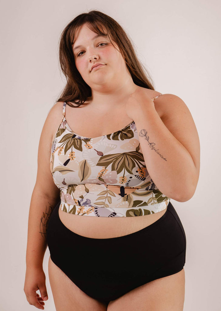 A woman in a Mango Mariposa Bralette Bikini Top by Mimi & August and black bottoms stands against a plain background. She has long hair and tattoos on her arms.