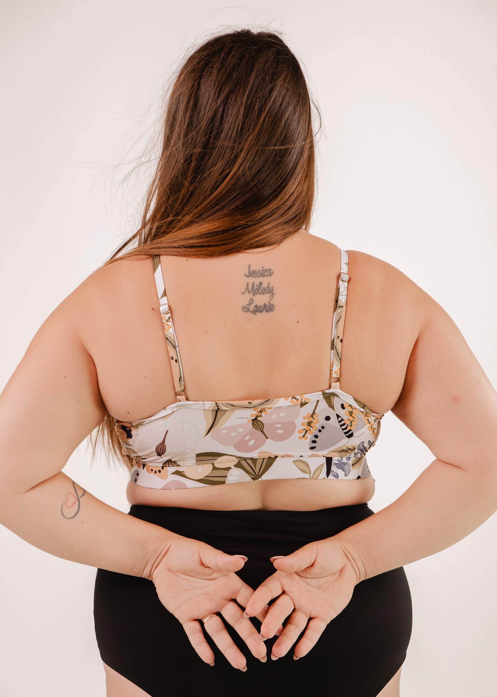 A person with long hair is seen from the back, wearing a Mimi & August Mango Mariposa Bralette Bikini Top adorned with a butterfly and floral print and black bottoms. Their hands are clasped behind their back, revealing a tattoo with names on their upper back.