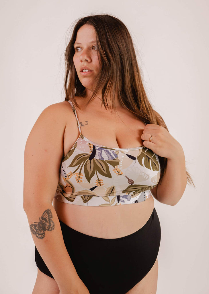 Woman with long hair wearing a Mimi & August Mango Mariposa Bralette Bikini Top and black bottoms. She has a tattoo of a butterfly on her upper arm and stands against a plain background, perfectly complementing the butterfly and floral print of her top.