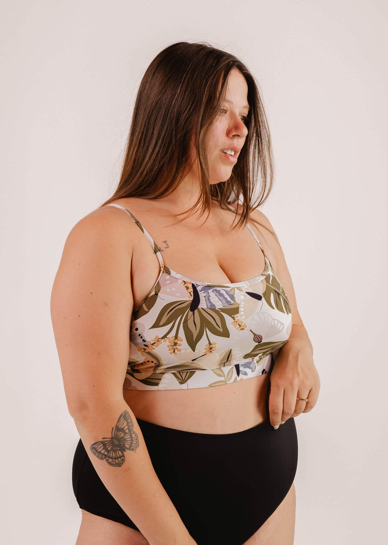 Une femme vêtue d'un haut de bikini Mimi & August Mango Mariposa Bralette et d'un short noir se tient debout sur un fond uni. Le haut de bikini, orné d'un papillon et d'un imprimé floral, offre un excellent soutien. Un papillon est tatoué sur son avant-bras gauche.