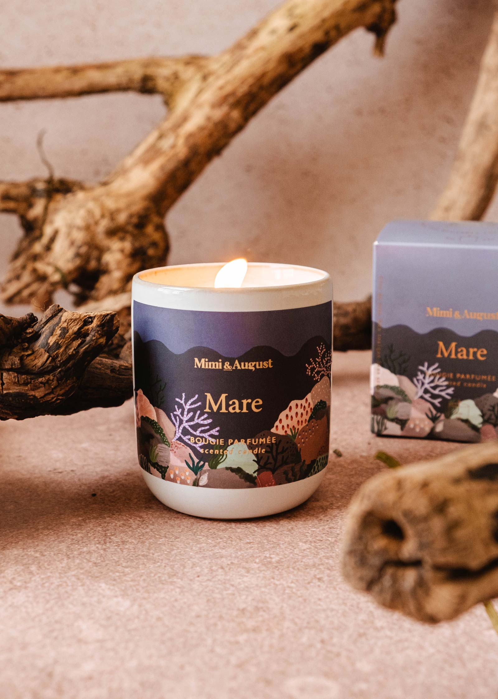 A lit "Mare - Reusable Candle" by Mimi & August sits on a surface with wooden branches in the background, and an unopened reusable candle box nearby.