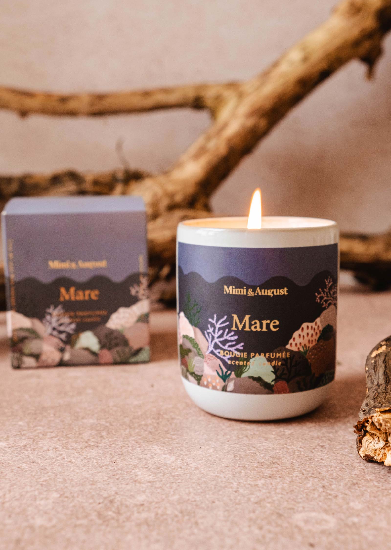 A lit "Mare - Reusable Candle" by Mimi & August, renowned for its calming qualities and ocean scent, placed on a surface with driftwood in the background. A matching product box is shown beside it.