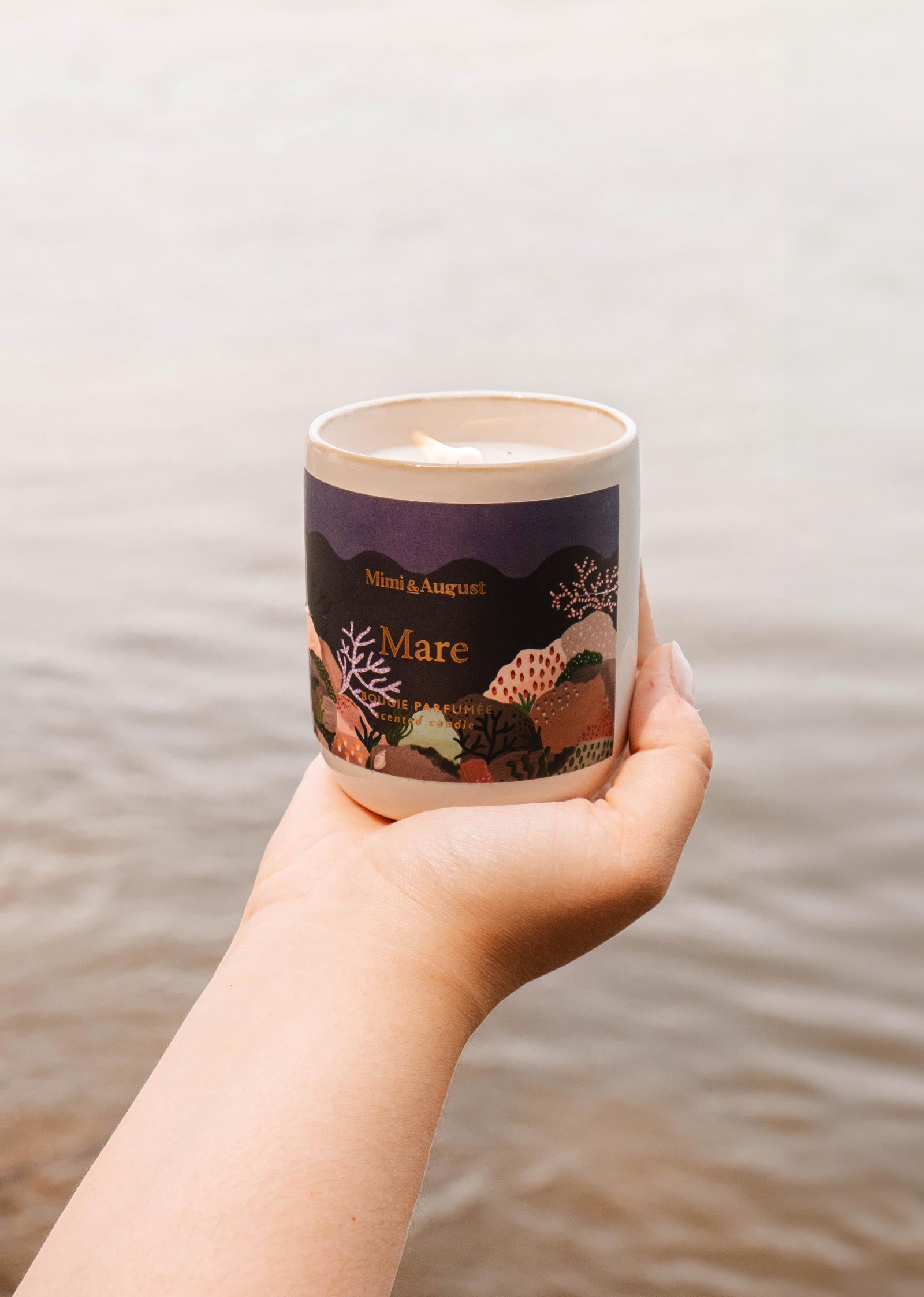 A hand holding a reusable candle with a dark-colored label that reads "Mare - Mimi & August." The background shows a body of water, hinting at its ocean scent and calming qualities.