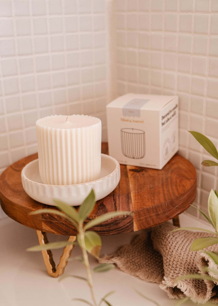 A white ribbed candle sits on a ceramic dish on a wooden stand, with a Candle Refill - Mare by Mimi & August nearby. Greenery and a textured fabric are also in the scene, set against a tiled backdrop. The ambiance is reminiscent of sea salt and ocean water, enhanced by eco-friendly candle refills.