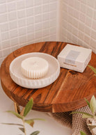 A round, white candle lies on a white dish on a wooden table. Next to it is the candle's packaging labeled "Candle Refill - Mare" by Mimi & August, featuring eco-friendly candle refills. The background showcases white tiled walls and some greenery, evoking the refreshing essence of ocean water.