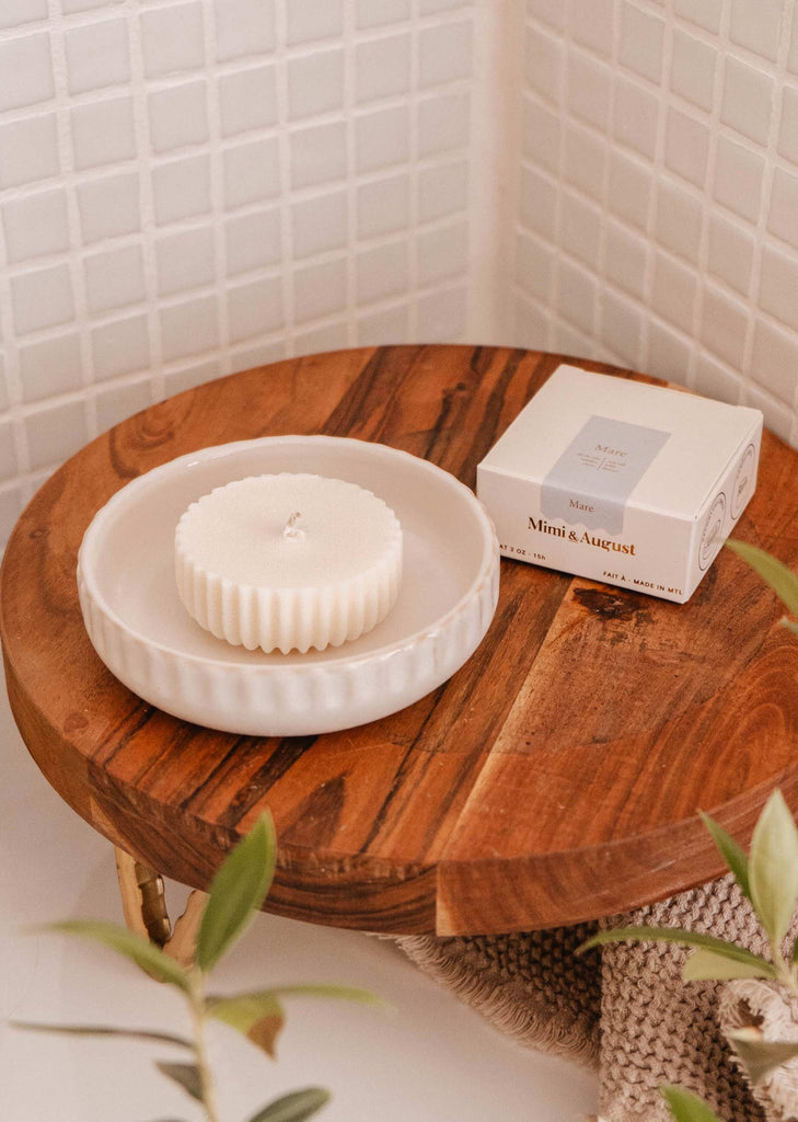 Une bougie ronde et blanche est posée sur un plat blanc sur une table en bois. À côté se trouve l'emballage de la bougie, étiqueté "Bougie rechargeable- Mare" par Mimi & August, qui propose des recharges de bougies respectueuses de l'environnement. L'arrière-plan présente des murs carrelés blancs et de la verdure, évoquant l'essence rafraîchissante de l'eau de mer.