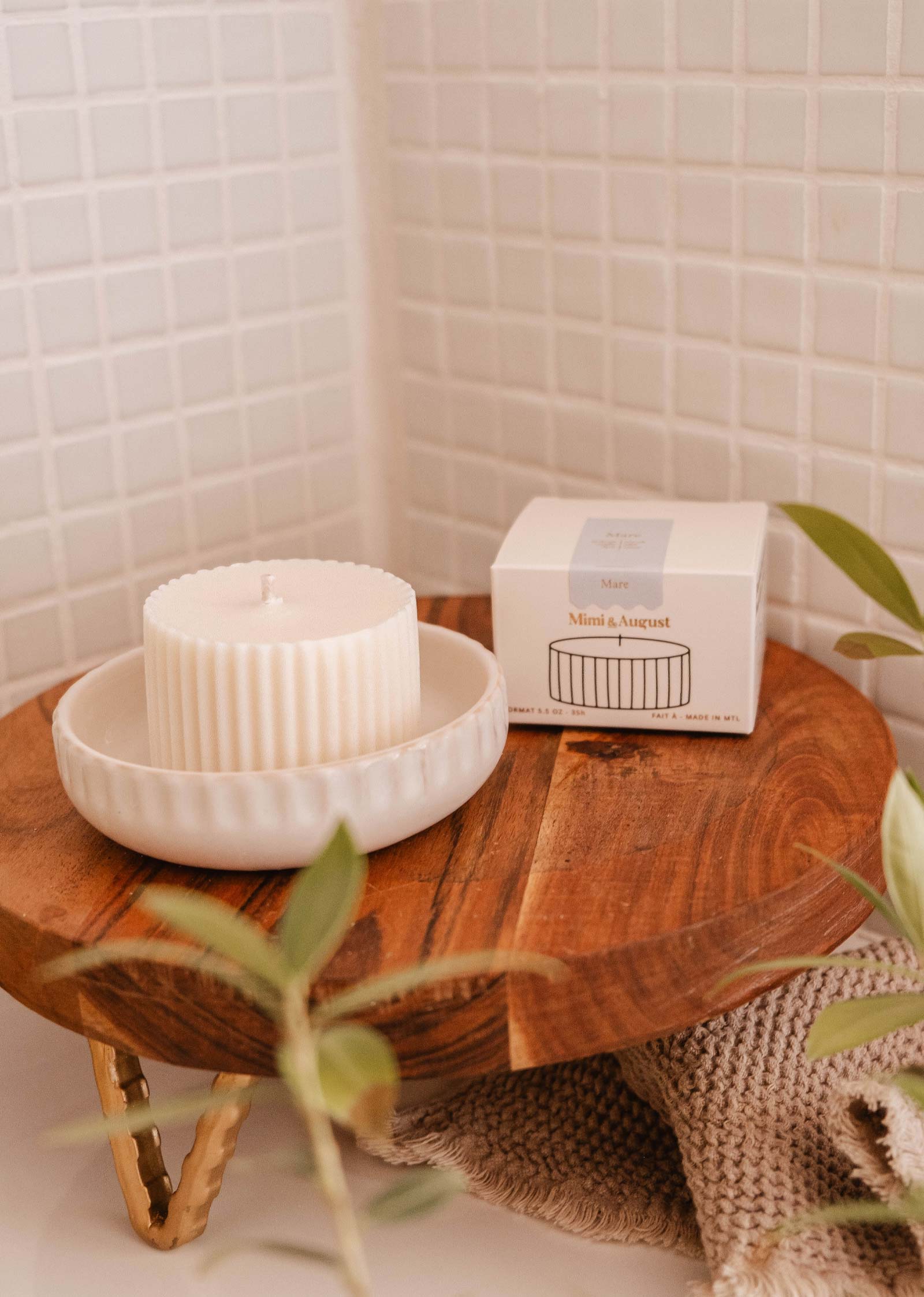 A fluted white candle refill infused with the essence of sea salt sits on a wooden tray in a ceramic dish beside a matching box labeled "Mimi & August" on a tiled surface, with some green leaves in the foreground.