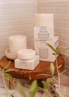 White ribbed candles displayed on a wooden stand, surrounded by green leaves against a tiled wall background. Some candles are placed in their packaging labeled "Mimi & August Candle Refill - Mare," and the air carries a hint of sea salt, reminiscent of ocean water.