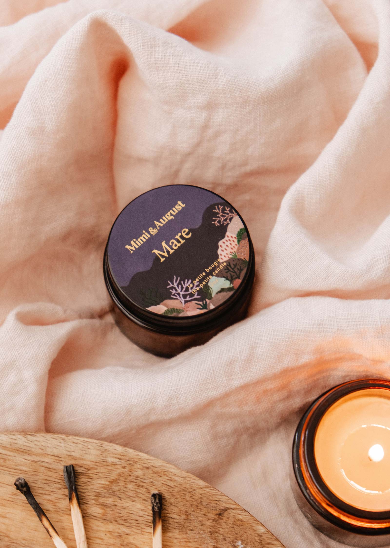 A Mimi & August Mare - 2 oz candle with an ocean scent rests on soft pink fabric beside a lit glass candle and burnt matches on wood, creating a tranquil vibe.