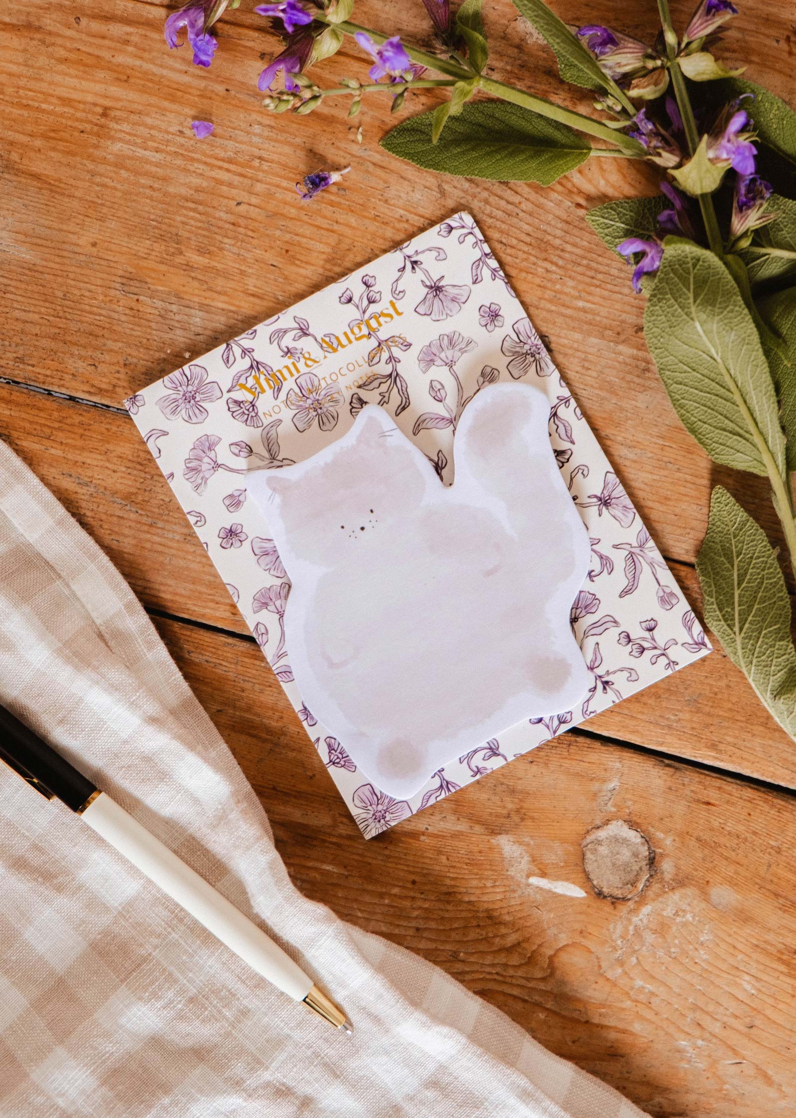 A Mimi & August "Meow - Sticky Notes" set, a pen, a checked cloth, and a floral notepad are arranged on wood with sprigs of purple flowers.