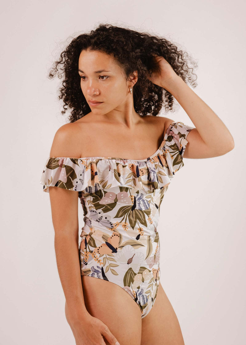 A person with curly hair wearing a cute Mimi & August Milano Mariposa One-Piece Swimsuit adorned with a butterfly and flower print stands against a plain background, holding their hair with one hand and looking to the side.