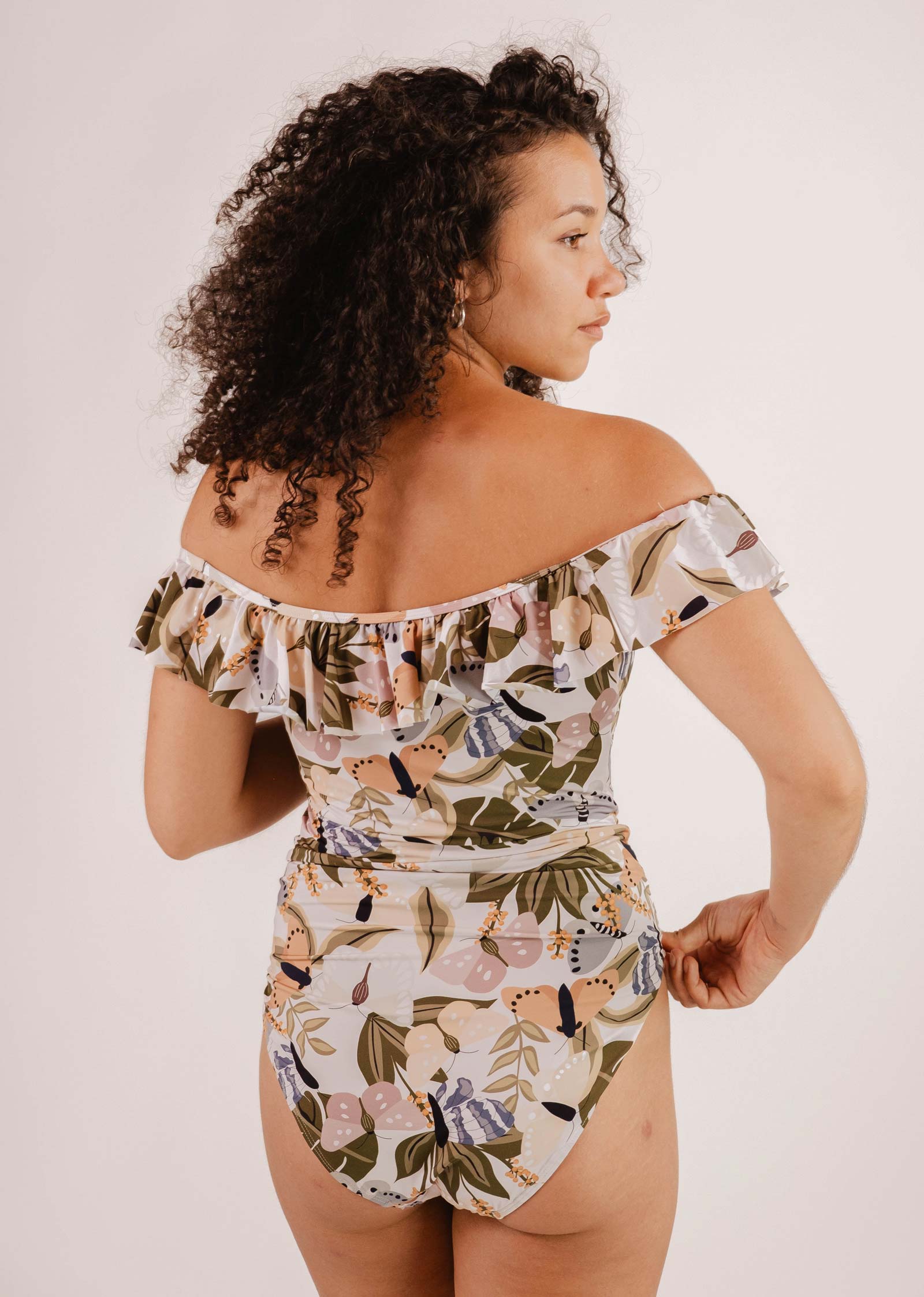 A person with curly hair stands sideways, donning a Mimi & August Milano Mariposa One-Piece Swimsuit showcasing a stunning butterfly and flower print.