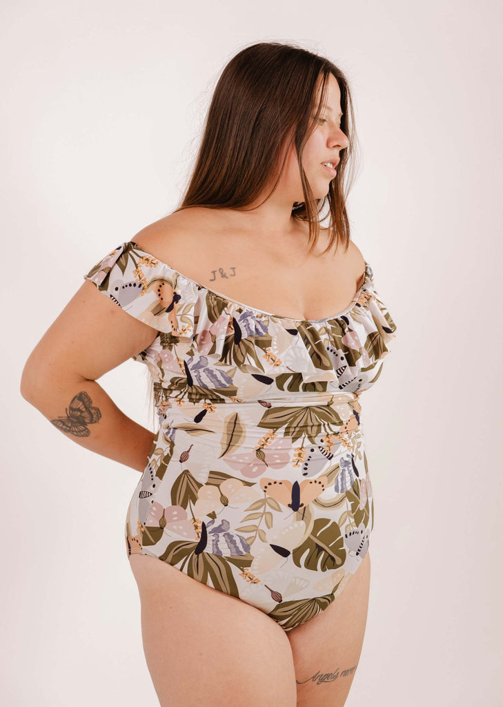 A woman in a Mimi & August Milano Mariposa One-Piece Swimsuit stands against a plain background. She has tattoos of butterflies and flowers on her upper arm and side.