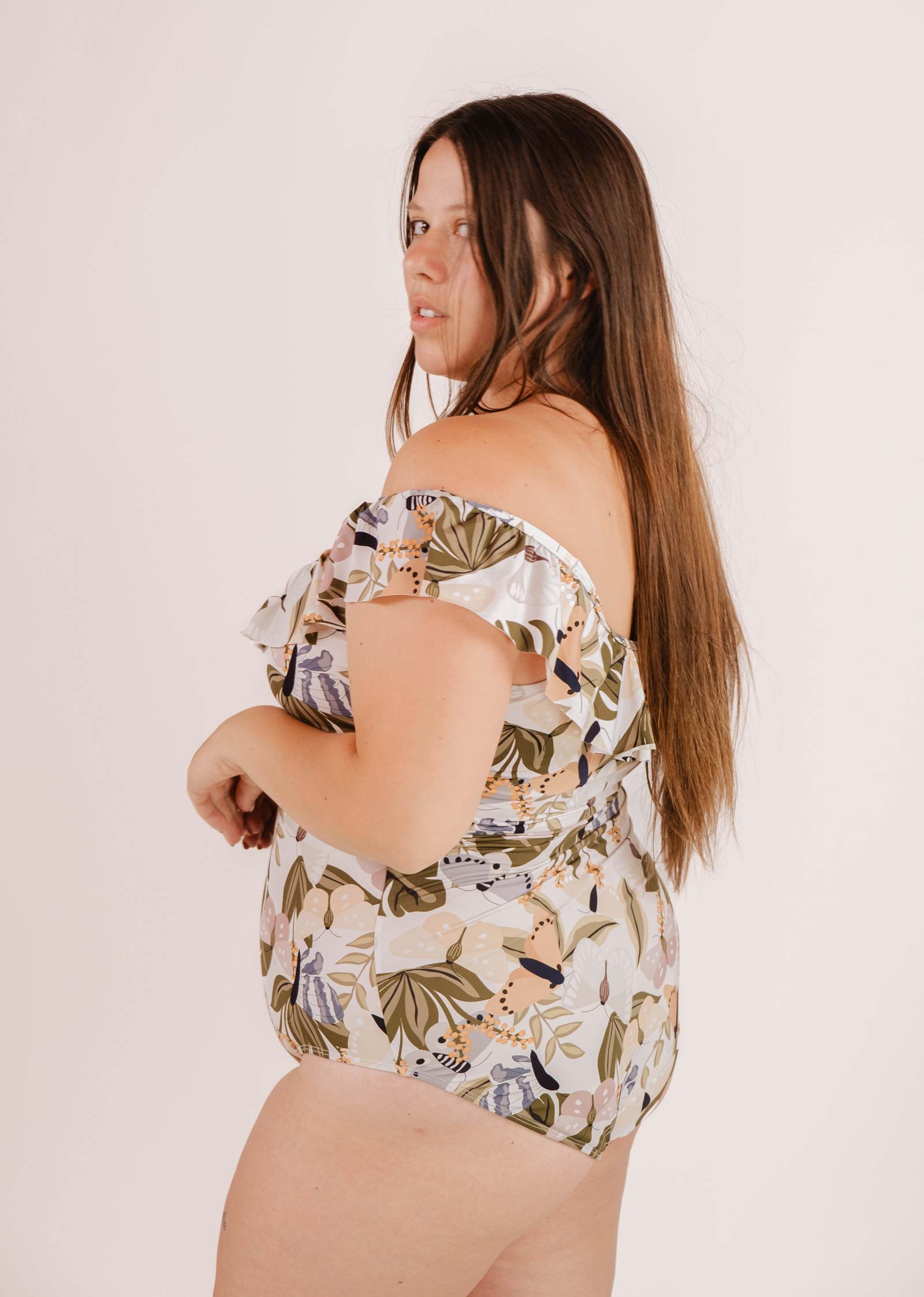 A person with long brown hair wearing an off-the-shoulder Milano Mariposa One-Piece Swimsuit by Mimi & August, featuring a delicate butterfly and flower print, poses looking back over their shoulder against a plain background.