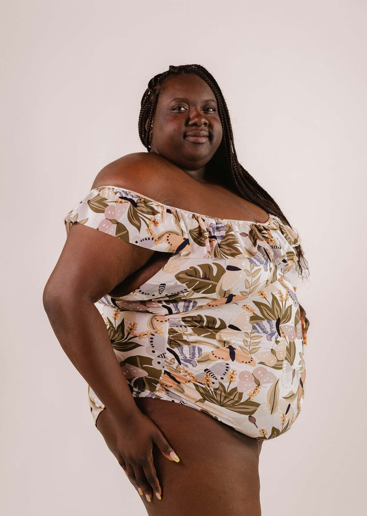 Person in a plus-size, floral off-shoulder Milano Mariposa One-Piece Swimsuit by Mimi & August poses confidently against a plain background.