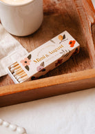 A Mimi & August Mimi Floral Matchbox - 30 Sticks is placed in a wooden tray next to a white cup.