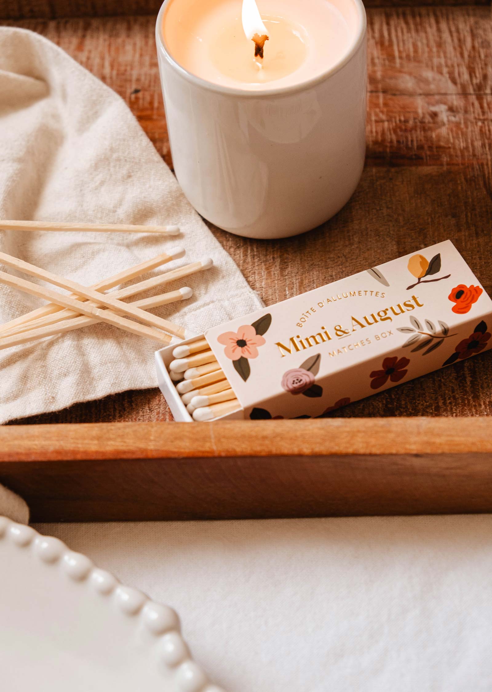 Une bougie allumée est posée sur une surface en bois à côté de la boîte d'allumettes Mimi & August Mimi Floral Matchbox - 30 Sticks, avec quelques-unes de ses allumettes écologiques éparpillées de manière fantaisiste sur un chiffon.