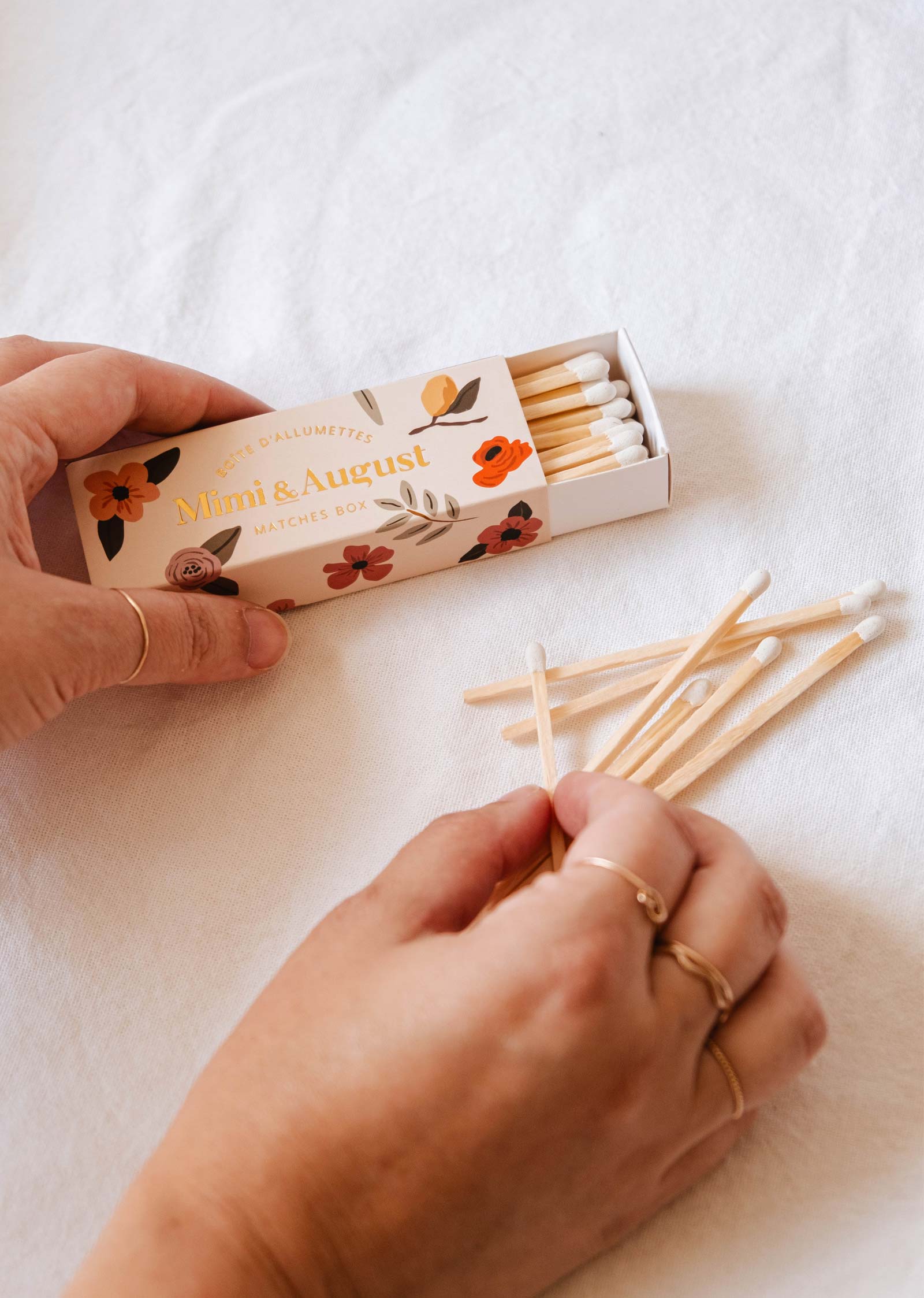 Hands holding and sorting eco-friendly matches next to an open Mimi & August Mimi Floral Matchbox - 30 Sticks with a whimsical design, all set against a white cloth background.