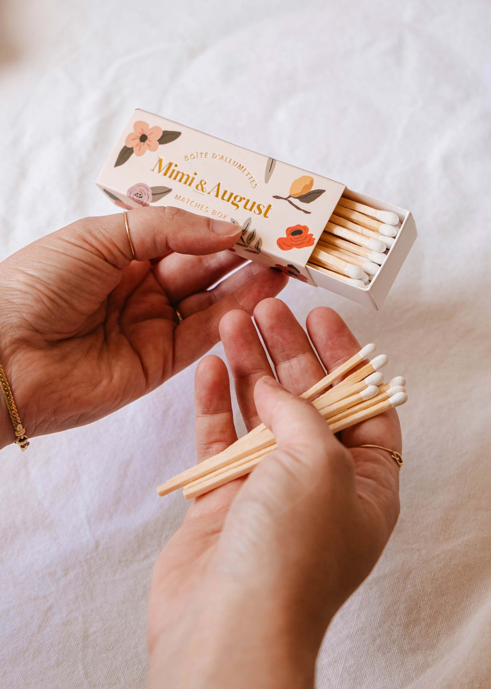 Two hands holding and arranging eco-friendly matches from a small, decorative matchbox labeled "Mimi Floral Matchbox - 30 Sticks by Mimi & August" on a white cloth surface.