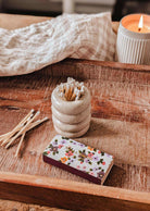 A wooden tray holds a lit candle, a ceramic match holder with decorative matches, several loose matches, and the Mimi Floral Matchbox - 30 Sticks by Mimi & August. A checked cloth is draped in the background.