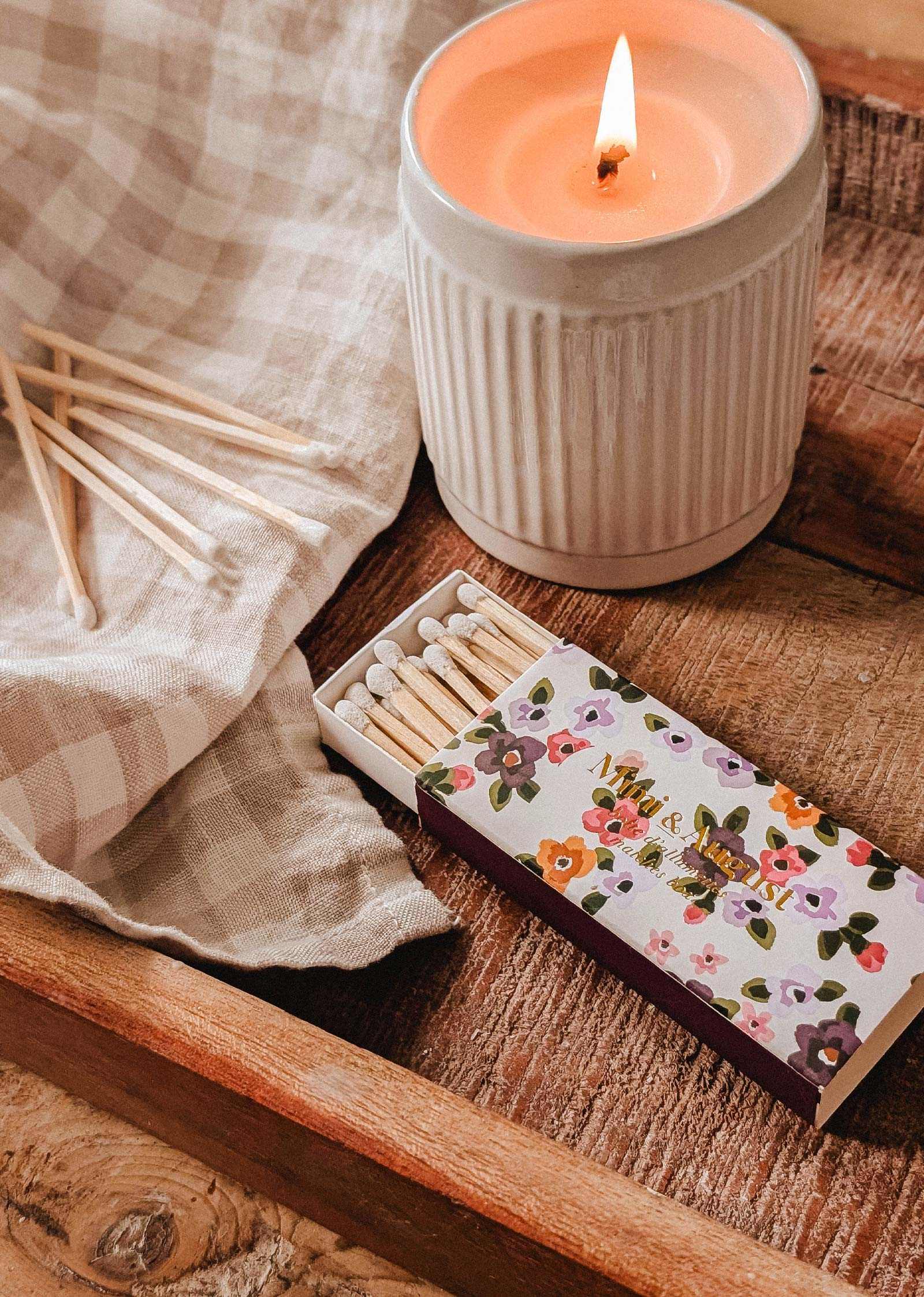 A lit candle in a ceramic holder rests on a wooden tray alongside a Mimi & August Mimi Floral Matchbox (30 sticks), several loose matches, and a checkered cloth.