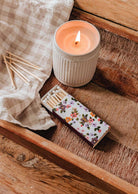 A lit candle in a ribbed white holder sits on a wooden tray beside the Mimi Floral Matchbox - 30 Sticks by Mimi & August, decorative matches, and a beige checkered cloth.