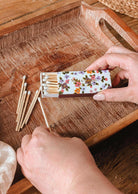 A person holds the Mimi & August Mimi Floral Matchbox - 30 Sticks and takes out a decorative, eco-friendly match, with several more scattered on a wooden tray.
