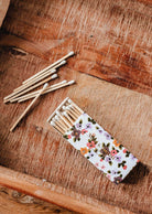 The Mimi & August Mimi Floral Matchbox - 30 Sticks, filled with eco-friendly matches, sits on a wooden surface with several matches scattered nearby.