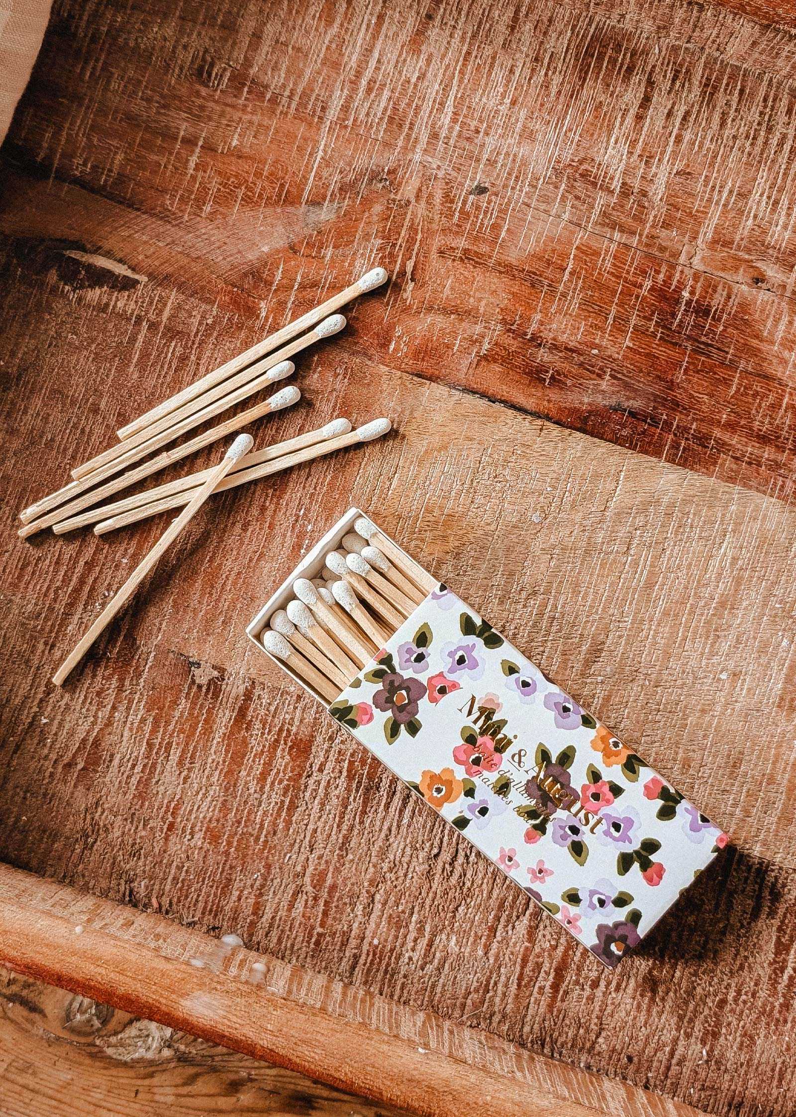 The Mimi & August Mimi Floral Matchbox - 30 Sticks, filled with eco-friendly matches, sits on a wooden surface with several matches scattered nearby.