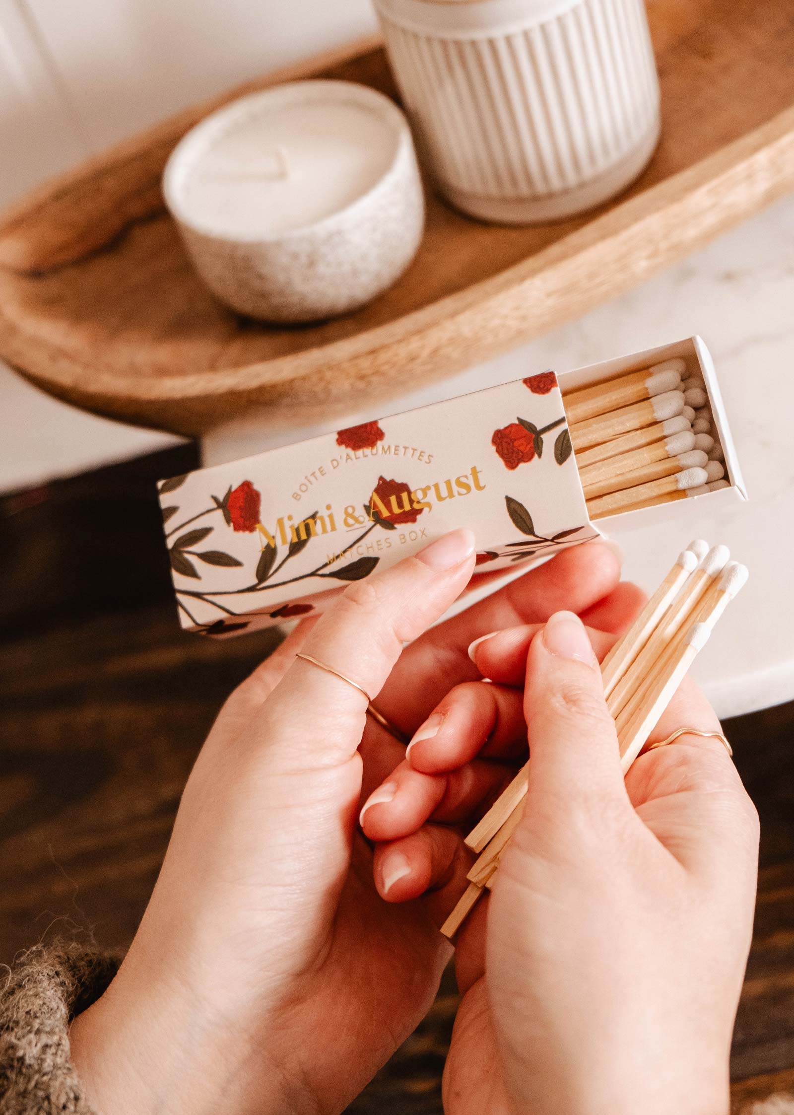 Des mains délicates tiennent une boîte d'allumettes écologique de Mimi & August étiquetée "Boîte d'allumettes florales Mimi - 30 bâtonnets", ornée de fleurs rouges. Une bougie vacille doucement sur un plateau en bois en arrière-plan, créant une scène sereine.