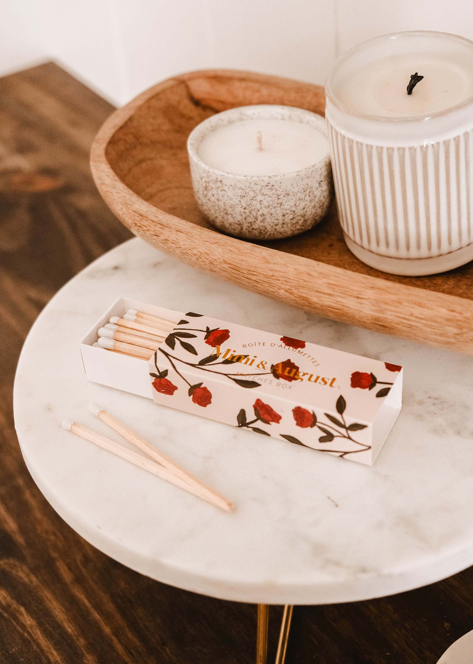 A Mimi Floral Matchbox from Mimi & August, featuring 30 eco-friendly sticks, sits on a marble table next to two lit candles in a wooden tray.