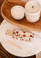 An open Mimi Floral Matchbox from Mimi & August, featuring an elegant red and white floral design, rests on a marble table near two lit candles on a wooden tray.