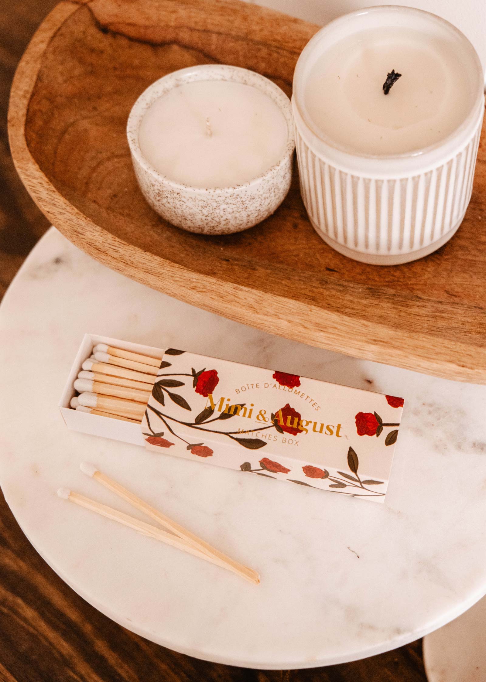 Une boîte d'allumettes ouverte Mimi Floral de Mimi & August, ornée d'un élégant motif floral rouge et blanc, repose sur une table en marbre près de deux bougies allumées sur un plateau en bois.