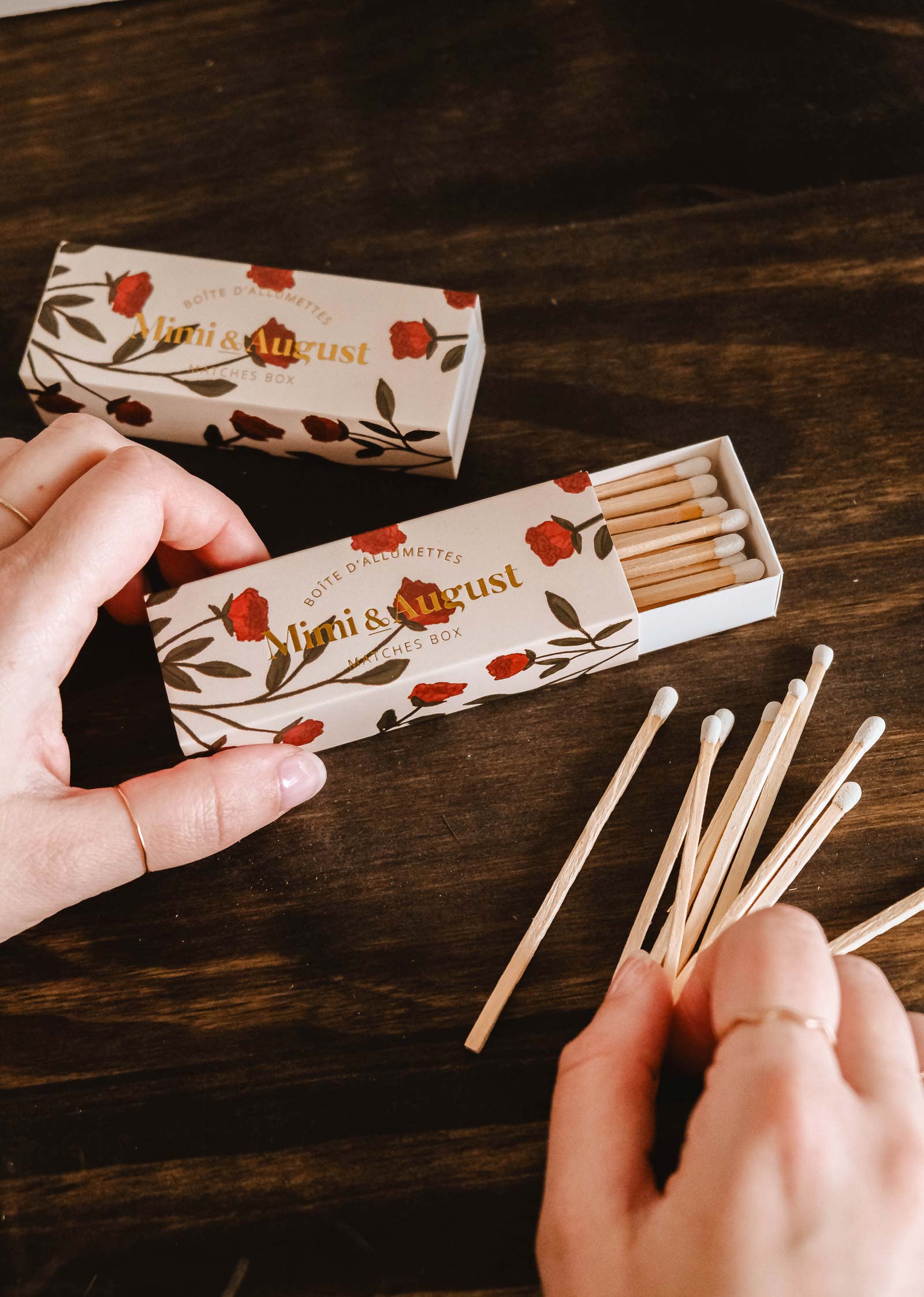 Les mains sélectionnent délicatement les allumettes d'une boîte d'allumettes Mimi & August ouverte, ornée d'un motif floral sur une surface en bois foncé, mettant en valeur l'élégante boîte d'allumettes Mimi Floral - 30 bâtonnets, fabriquée à partir de matériaux écologiques.