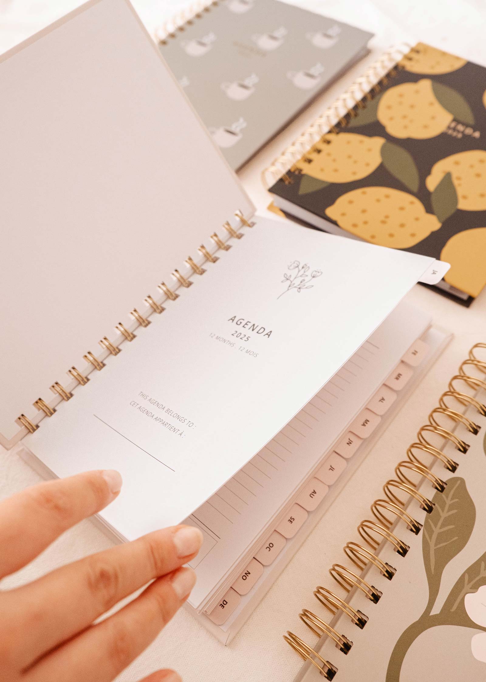 Une main feuillette l'agenda Mimi & August Champs de fleurs avec sa solide reliure à spirale sur une table, entouré d'autres carnets fermés ornés de divers motifs de fruits, chacun doté d'une couverture résistante.