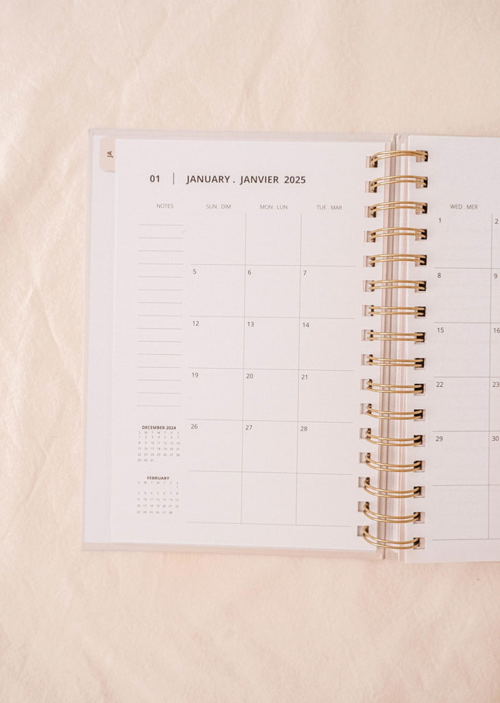 An open spiral-bound planner, the Mimi & August Lemon - Agenda 2025, lies on a light fabric surface. The January 2025 calendar is visible, adorned with a delightful hand-drawn lemon pattern.