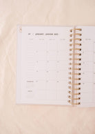 The Cafecito - Agenda 2025 planner by Mimi & August rests open on a light fabric surface, its spiral binding revealing the January calendar. Designed for coffee enthusiasts, it ensures an organized beginning to your caffeine-fueled year.