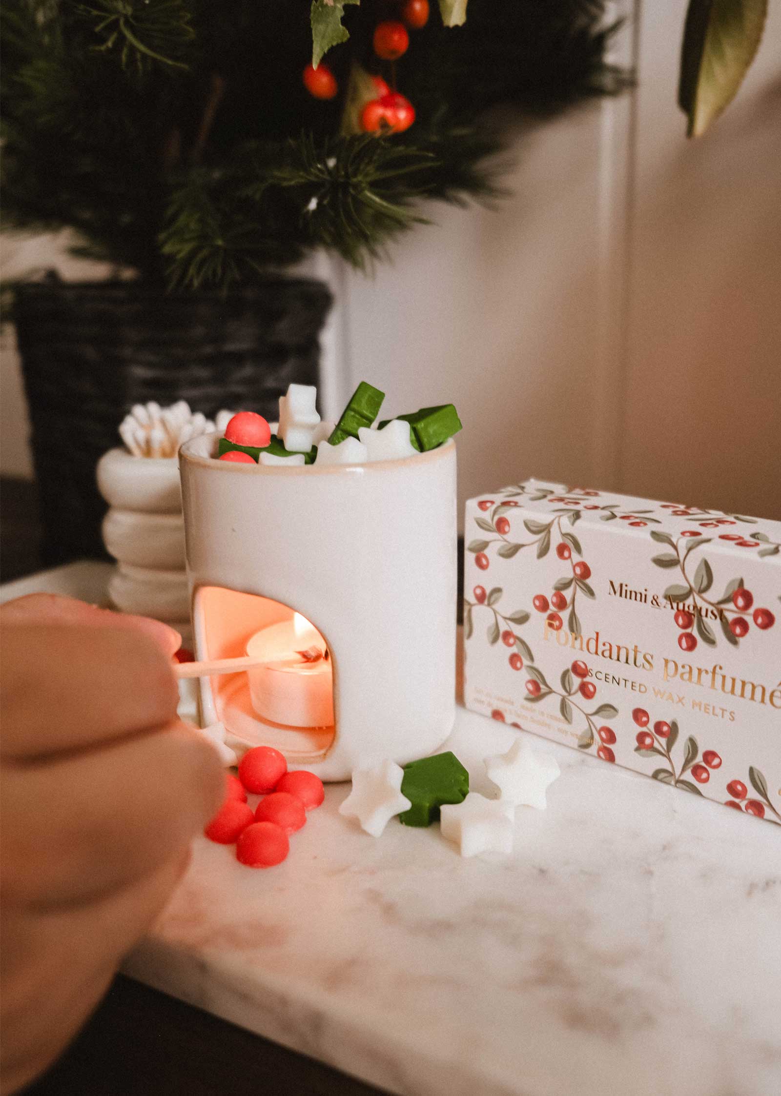A hand lights a tealight under the Mimi & August Mini Aroma Diffuser, surrounded by colorful wax melts, with a wax melts box and a potted plant in the background.