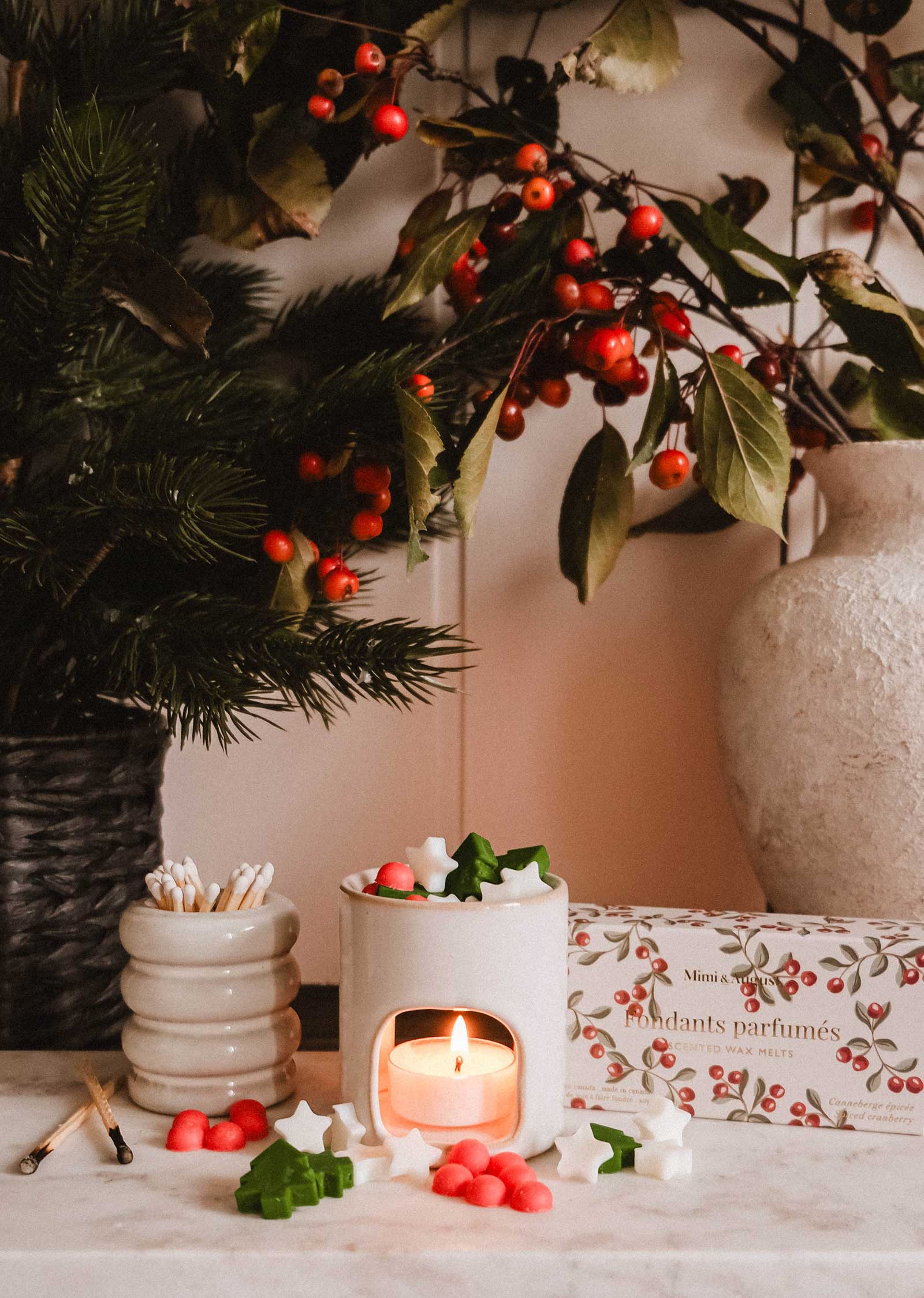 A lit white candle warmer with wax melts, matches, and a match holder sits on marble next to a floral arrangement, a decorative box, and the Mimi & August Mini Aroma Diffuser.