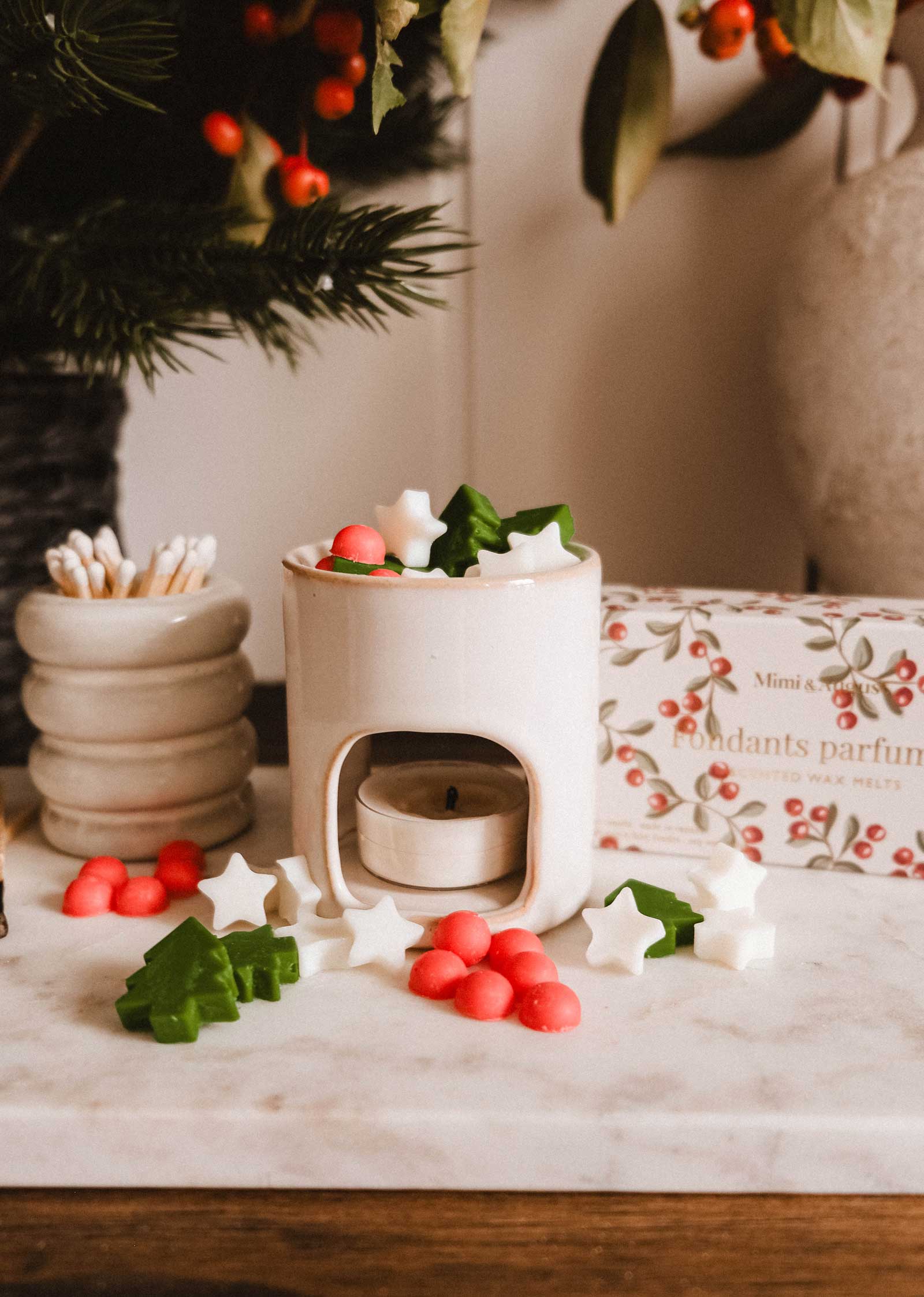 The Mimi & August Mini Aroma Diffuser, a white ceramic piece with a lit tealight, is surrounded by festive star, tree, and berry wax melts on a marble surface beside its box and a cotton swab container.