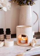 A lit tealight candle glows inside the Mini Aroma Diffuser by Mimi & August, surrounded by aromatherapy oils, wax melts, and dried flowers in a vase.