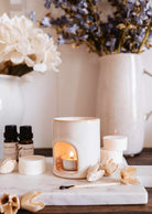 The Mini Aroma Diffuser by Mimi & August, a white ceramic piece with a lit tealight, sits on a marble tray with essential oils, wax melts, matches, and vases filled with decorative flowers.