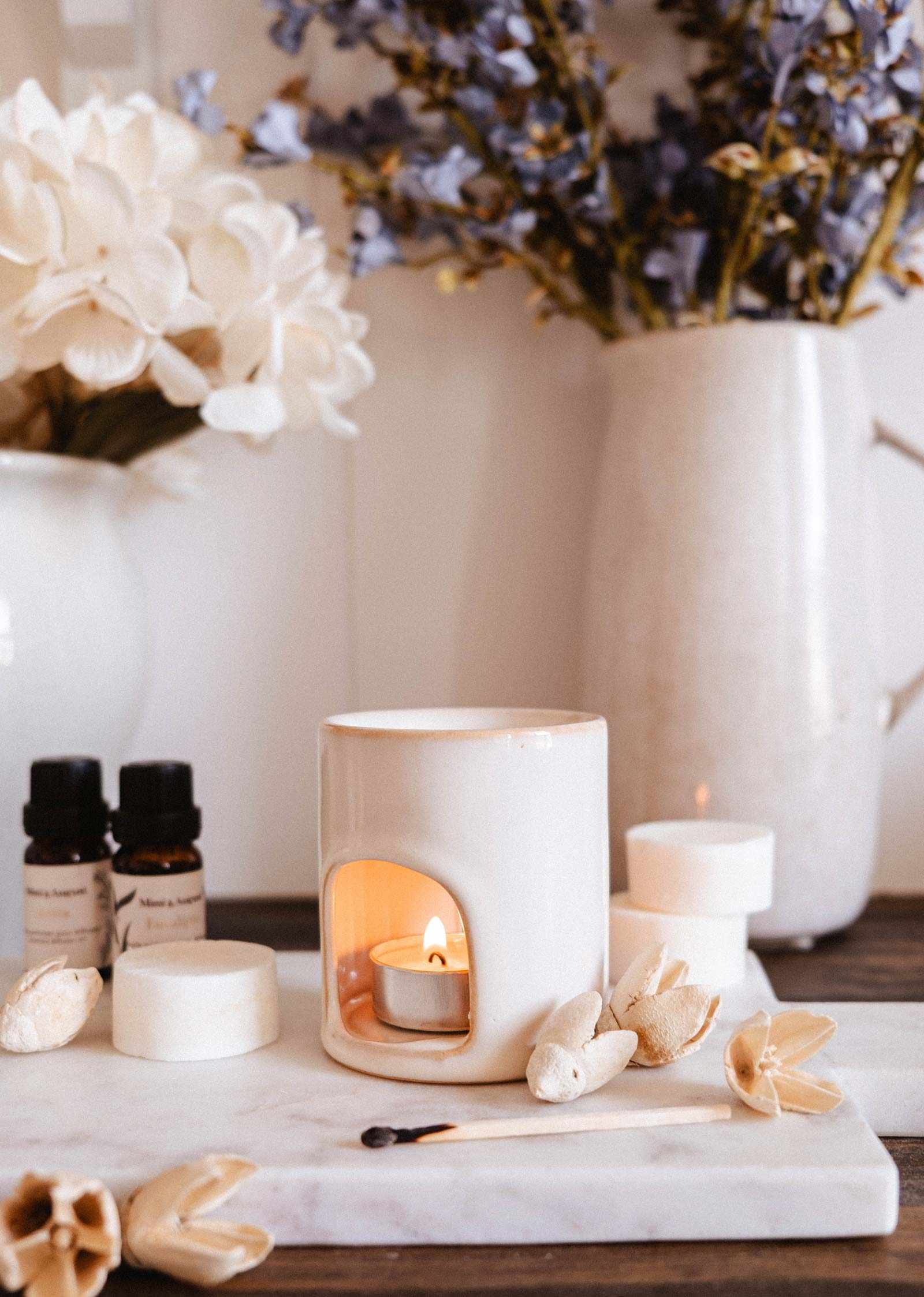 The Mini Aroma Diffuser by Mimi & August, a white ceramic piece with a lit tealight, sits on a marble tray with essential oils, wax melts, matches, and vases filled with decorative flowers.
