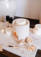The Mimi & August Mini Aroma Diffuser, a white ceramic piece with a candle inside, is displayed on marble alongside wax melts, dried flowers, essential oil bottles, and a used matchstick.